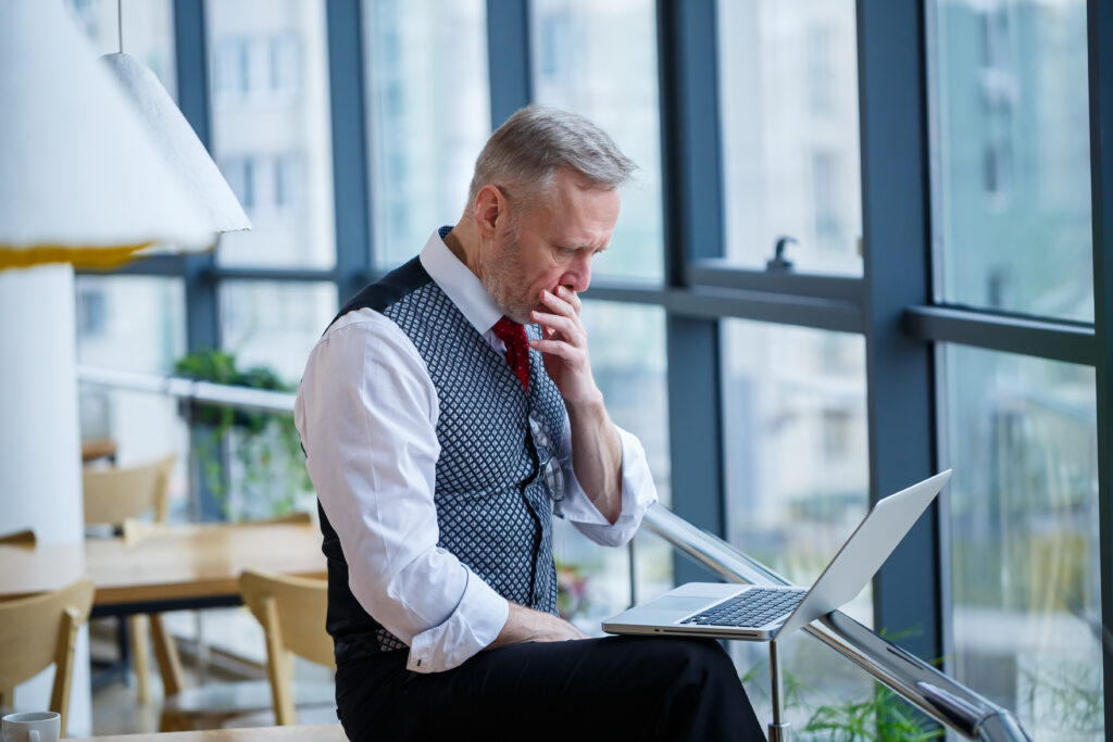 Adult male businessman, teacher, mentor working on a new project. Sits by a large window on the table. He looks at the laptop screen